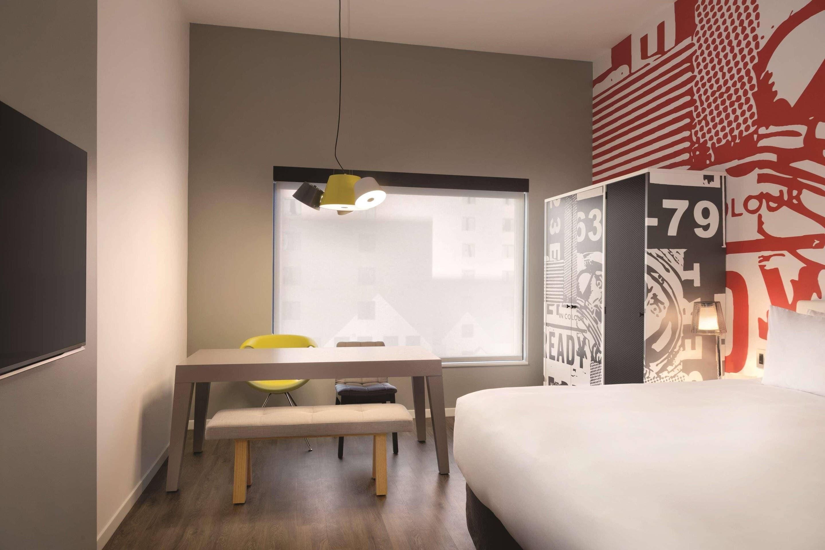Une chambre d’hôtel moderne, équipée d’un lit, d’un bureau avec une chaise, d’une fenêtre et d’un mur sur lequel figure un grand numéro.