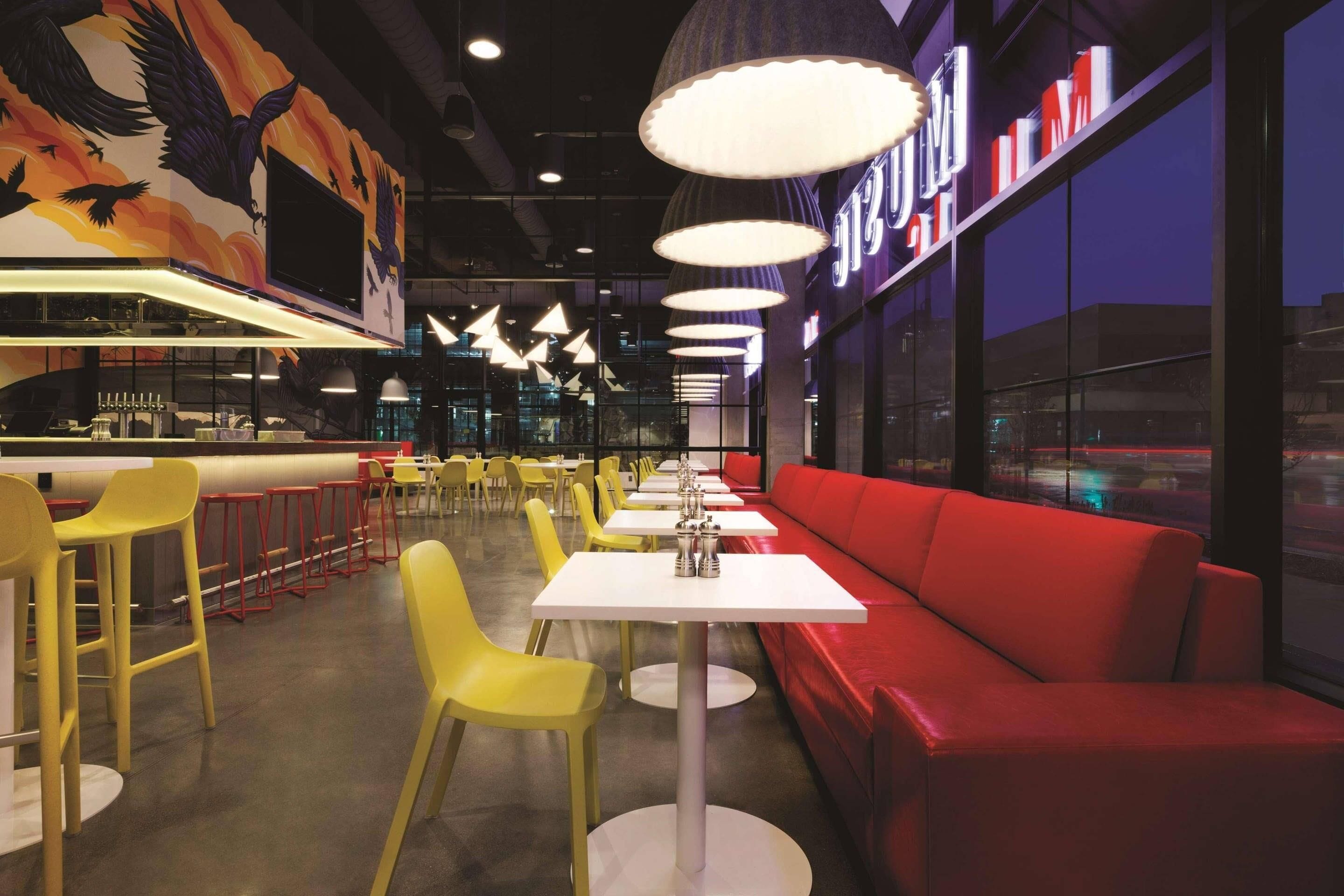 A restaurant interior with yellow chairs, white tables, red seating, and pendant lighting.