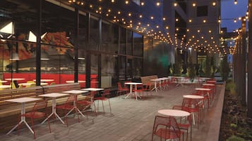 An outdoor seating area with tables and chairs, string lights overhead, and a view of a building with large windows.