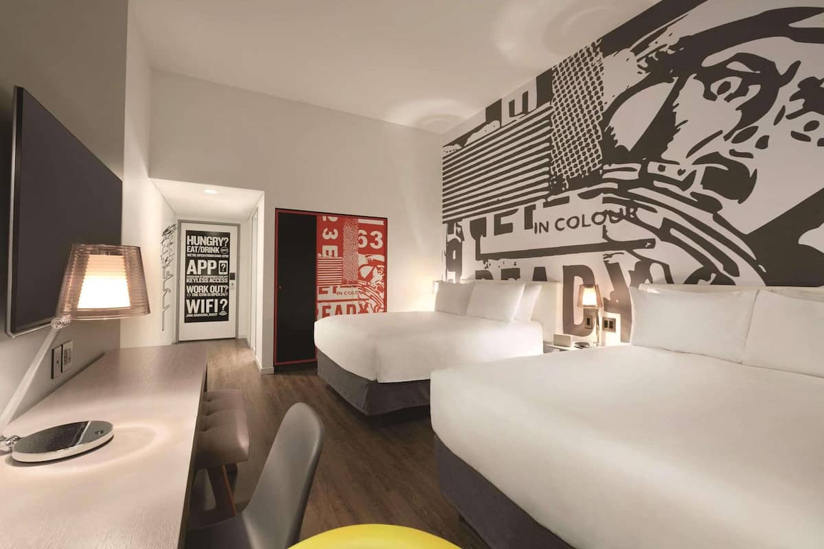 A hotel room with two beds, a desk with a lamp, and a large wall mural.