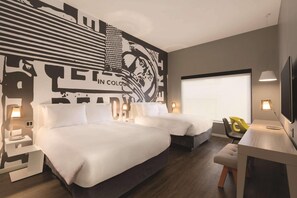 Habitación de hotel con dos camas, un escritorio con silla, un ventanal grande y un mural en la pared.