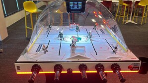 Una mesa de fútbol de mesa con marcador digital y luces LED, ubicada en una sala con sillas amarillas y una pantalla de proyección al fondo.