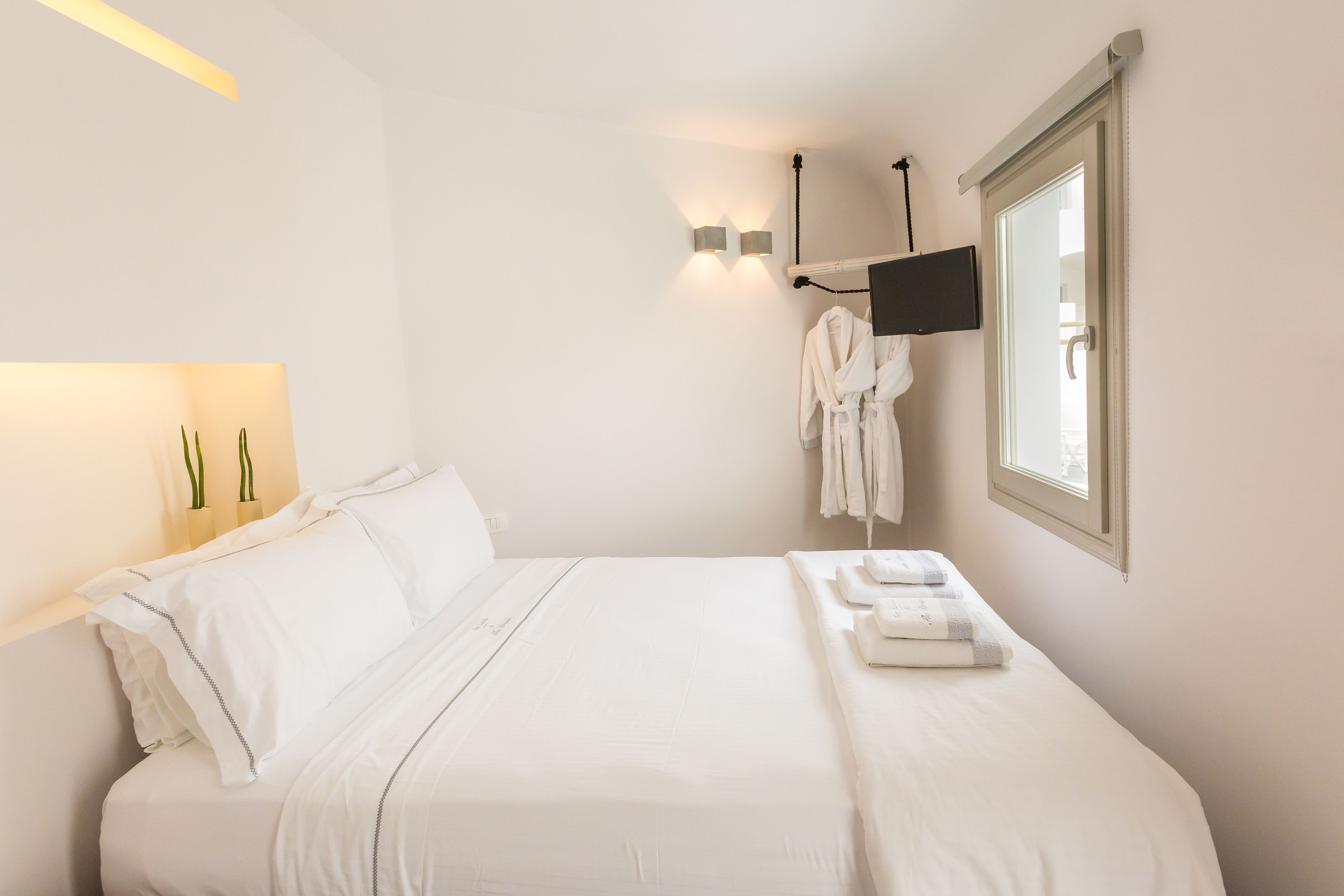 A neatly made bed with white linens, a flat-screen TV mounted on the wall, and a window with a view of the outside.