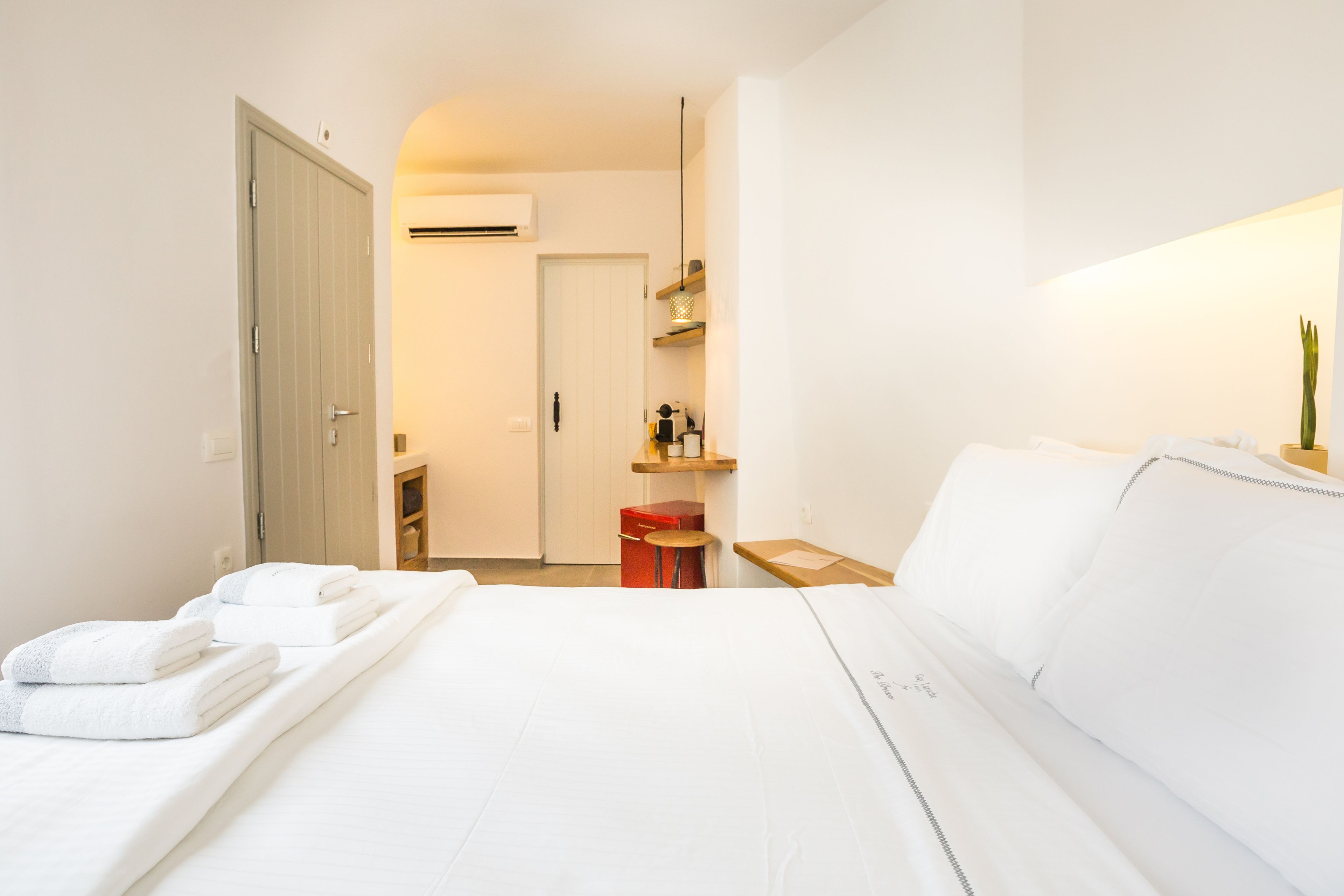 Een hotelkamer met een bed, een nachtkastje, een rode koffer, een klein krukje, een plank met toiletartikelen en een airconditioner.