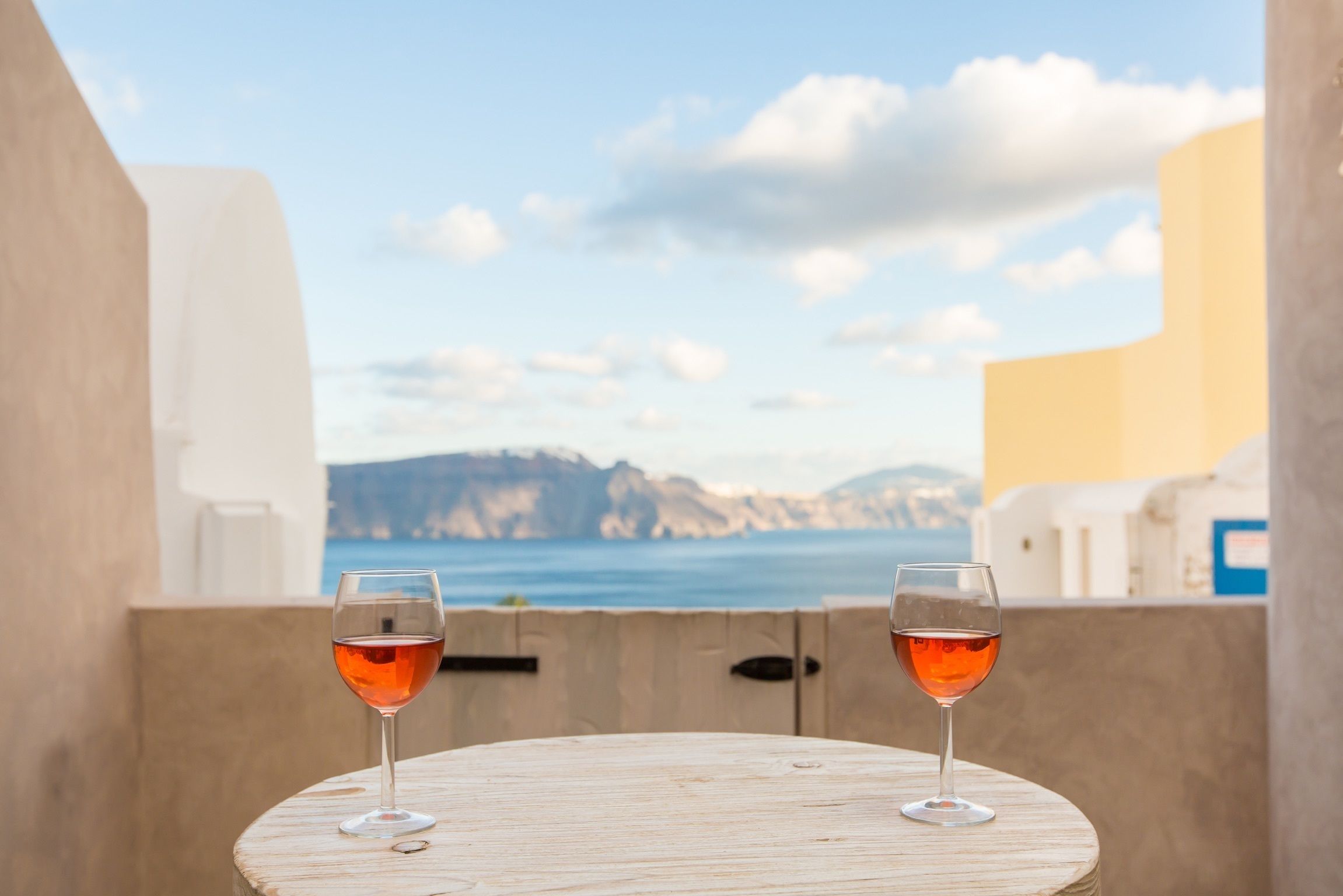 Two wine glasses on a round wooden table with a view of the sea and mountains in the background.