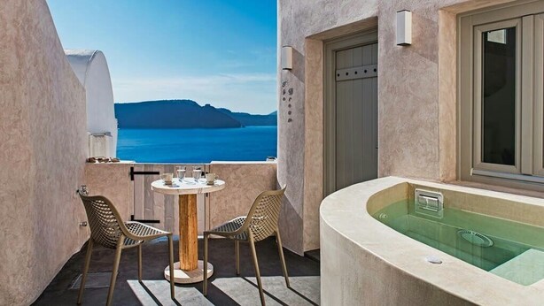 Un balcón con una mesa redonda y dos sillas, una bañera, y vistas al mar y a las montañas.