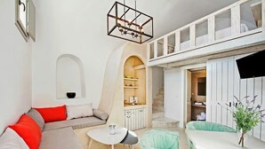 Una sala de estar moderna con un sofá, una mesa redonda y un sillón informal de color verde. Hay un mueble blanco con estantes y un ventanal pequeño.