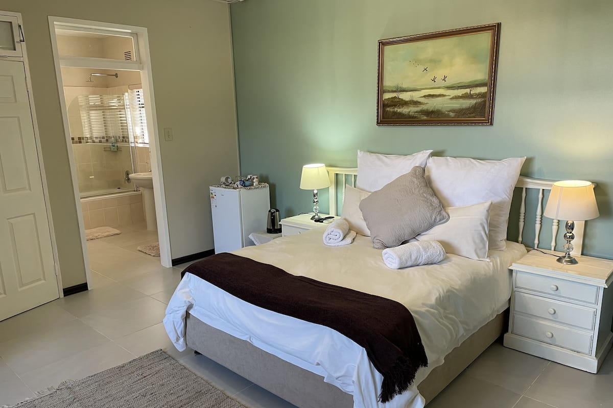 A neatly made bed with white linens and a brown throw blanket, a nightstand with a lamp, and a painting on the wall.