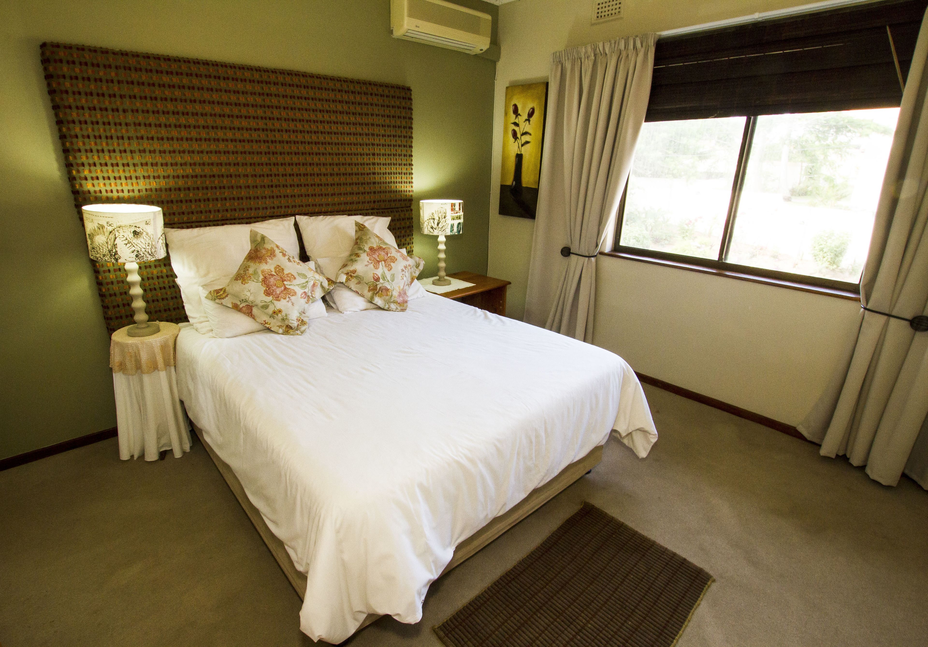 A neatly made bed with white linens and floral pillows, a bedside lamp, and a window with curtains.