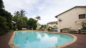 A rectangular swimming pool with a brick border, surrounded by lush greenery and a building with a beige exterior. - Eden Rose Manor (La Mercy)