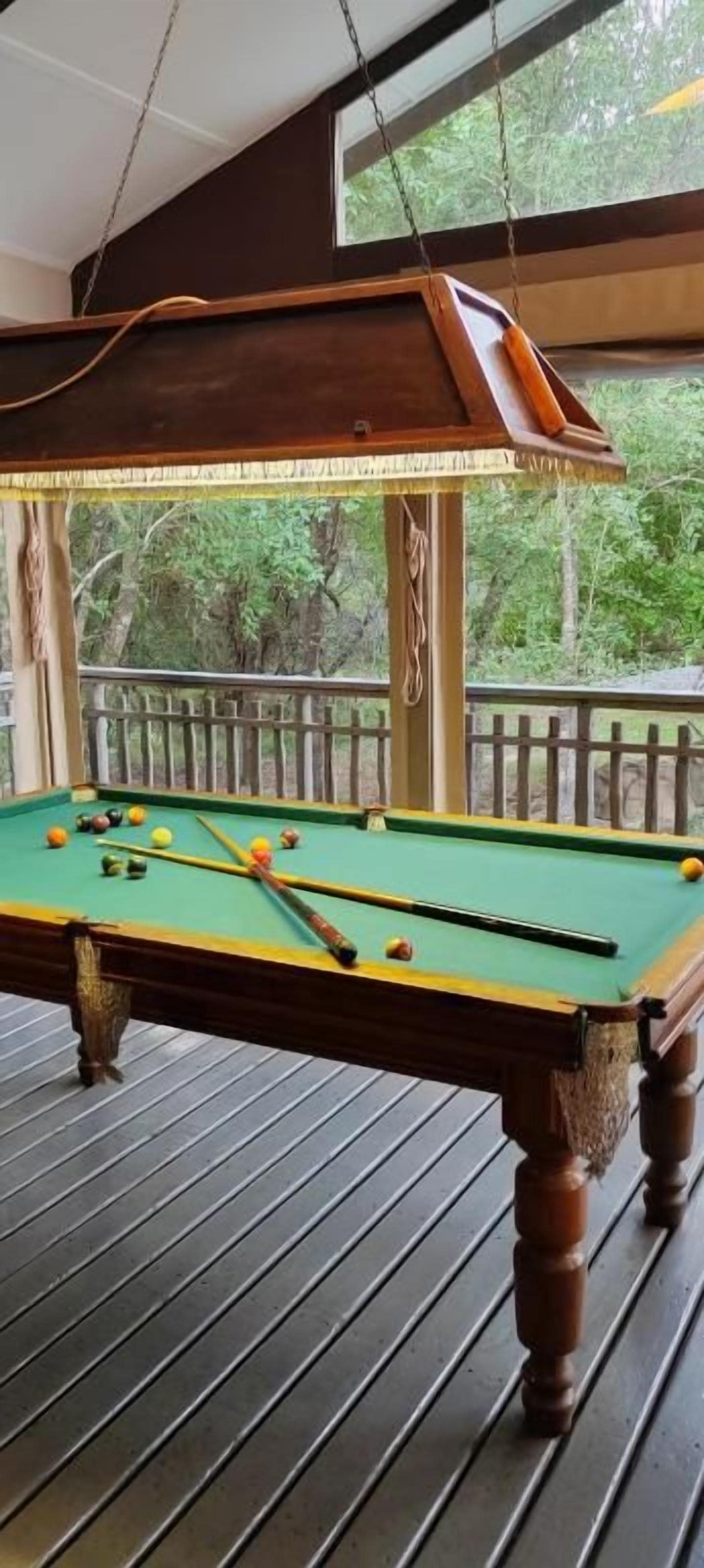 A pool table with balls on a wooden deck, a hanging lamp, and a view of trees through a window.