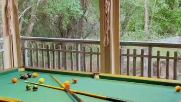 A pool table with balls on a wooden deck, a hanging lamp, and a view of trees through a window.