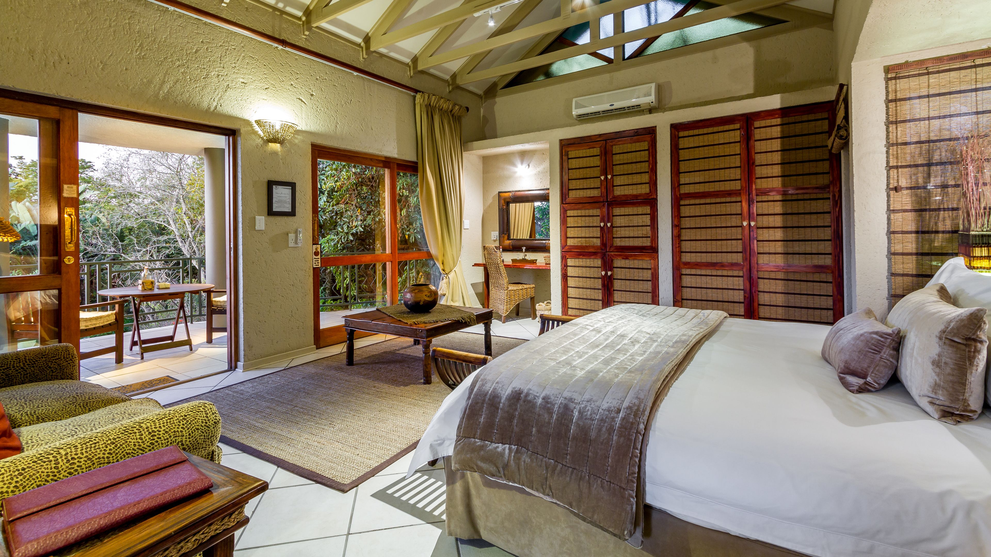 A bedroom with a bed, a desk, and a chair. There is a window with curtains, a door, and a view of greenery outside.