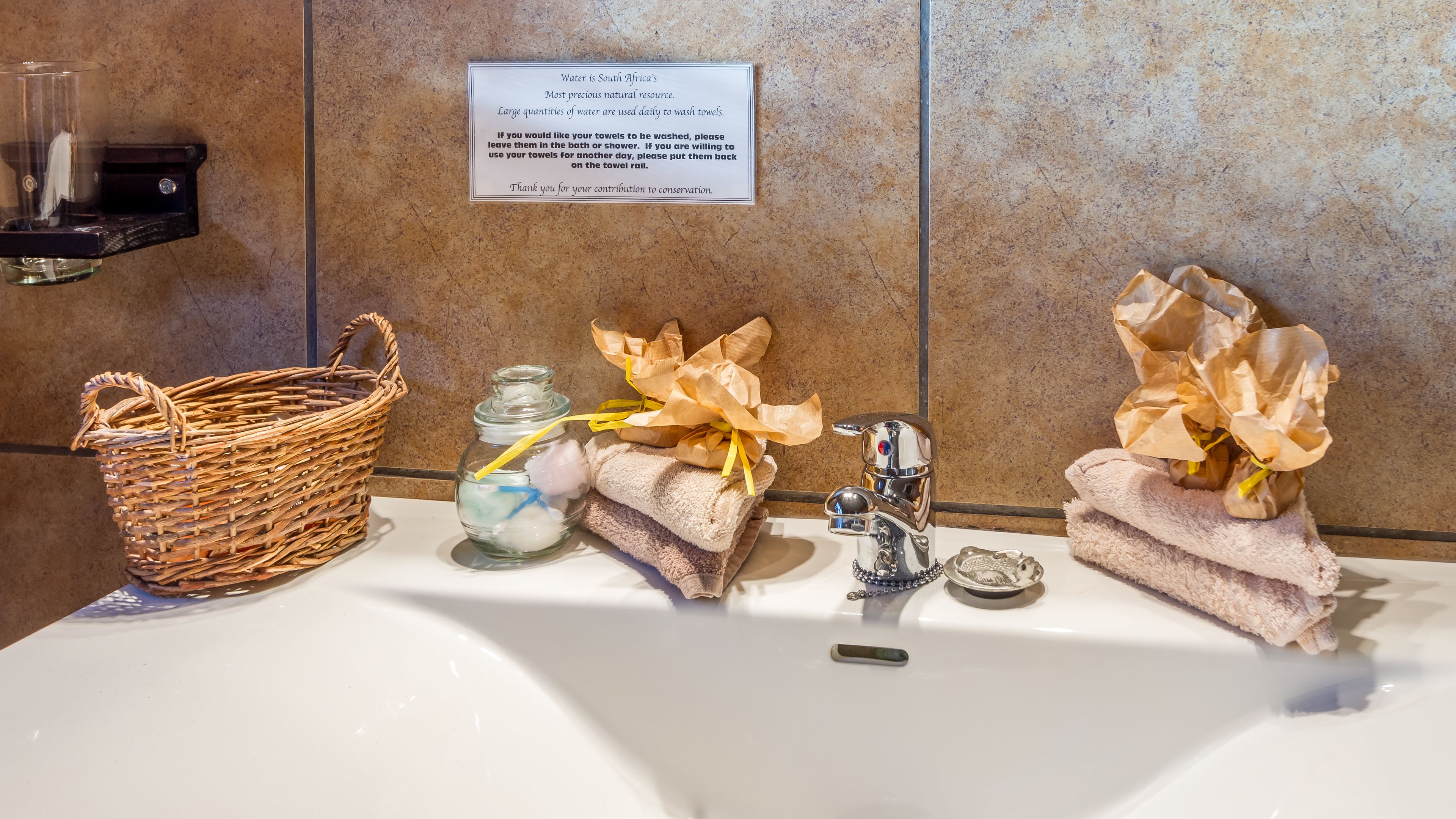 A bathroom sink with a towel rack, a glass jar with a yellow straw, and a sign about water conservation.