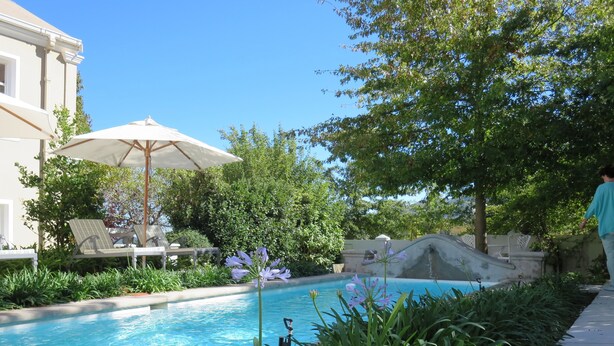 Une piscine dotée de chaises longues et d’un parasol, entourée de verdure et d’un bâtiment.