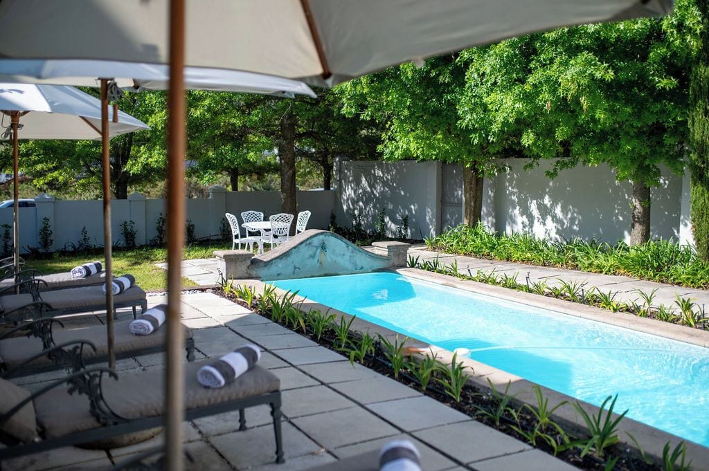 A swimming pool with lounge chairs and umbrellas, surrounded by greenery and a white wall.
