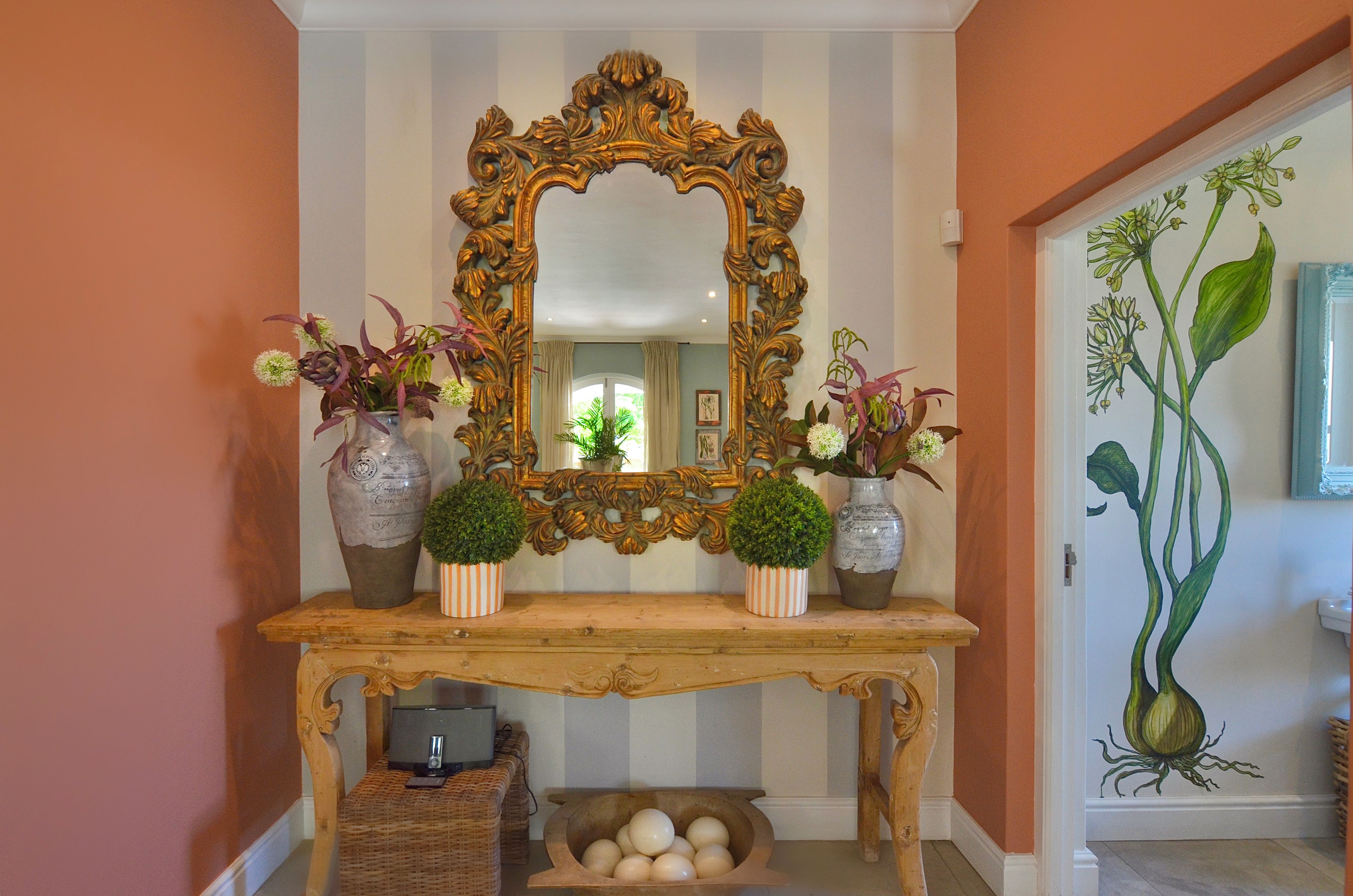Un couloir avec un miroir, deux vases ornés de plantes, un panier en osier et un mur décoré d’une illustration de plantes.