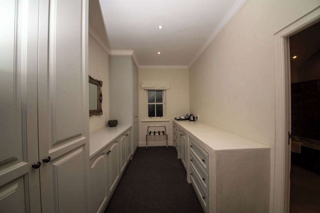 A narrow hallway with white walls and a window. There is a small bench and a coat rack near the window.
