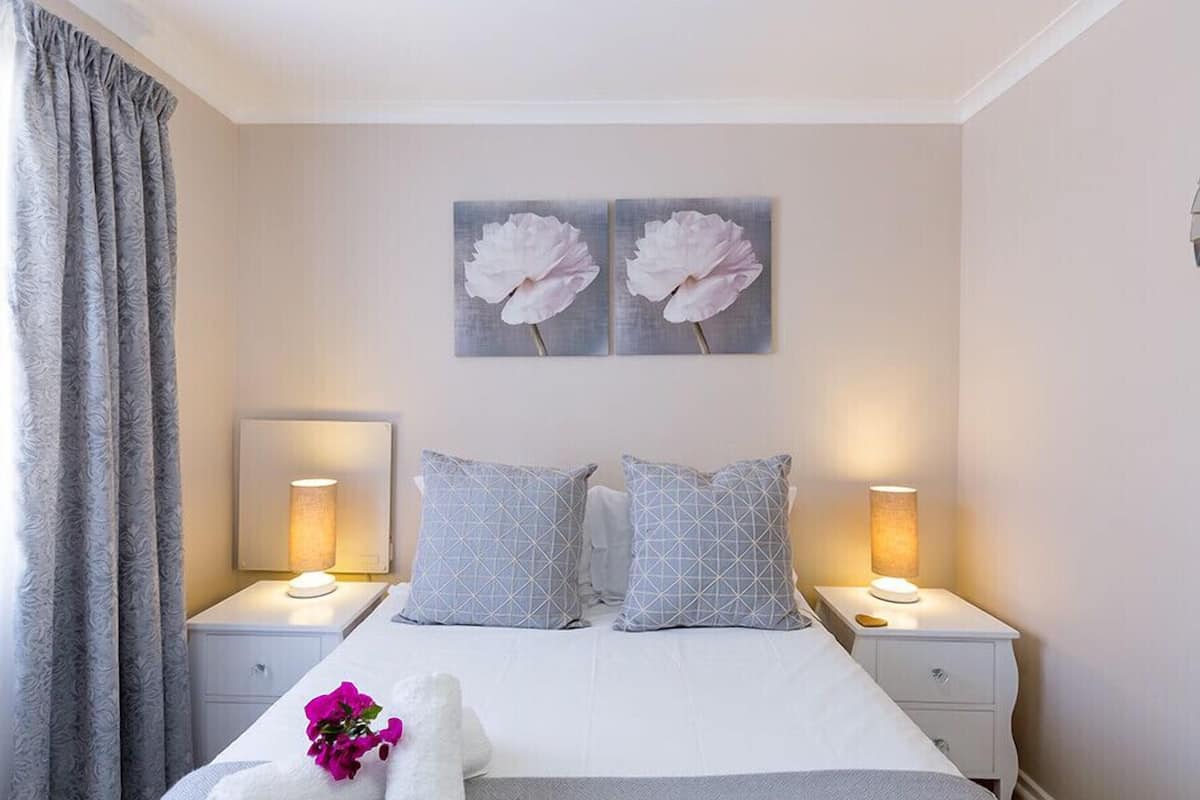 Quarto de hotel com cama, duas mesas de cabeceira com abajures, uma janela grande com cortinas e dois quadros de flores na parede.