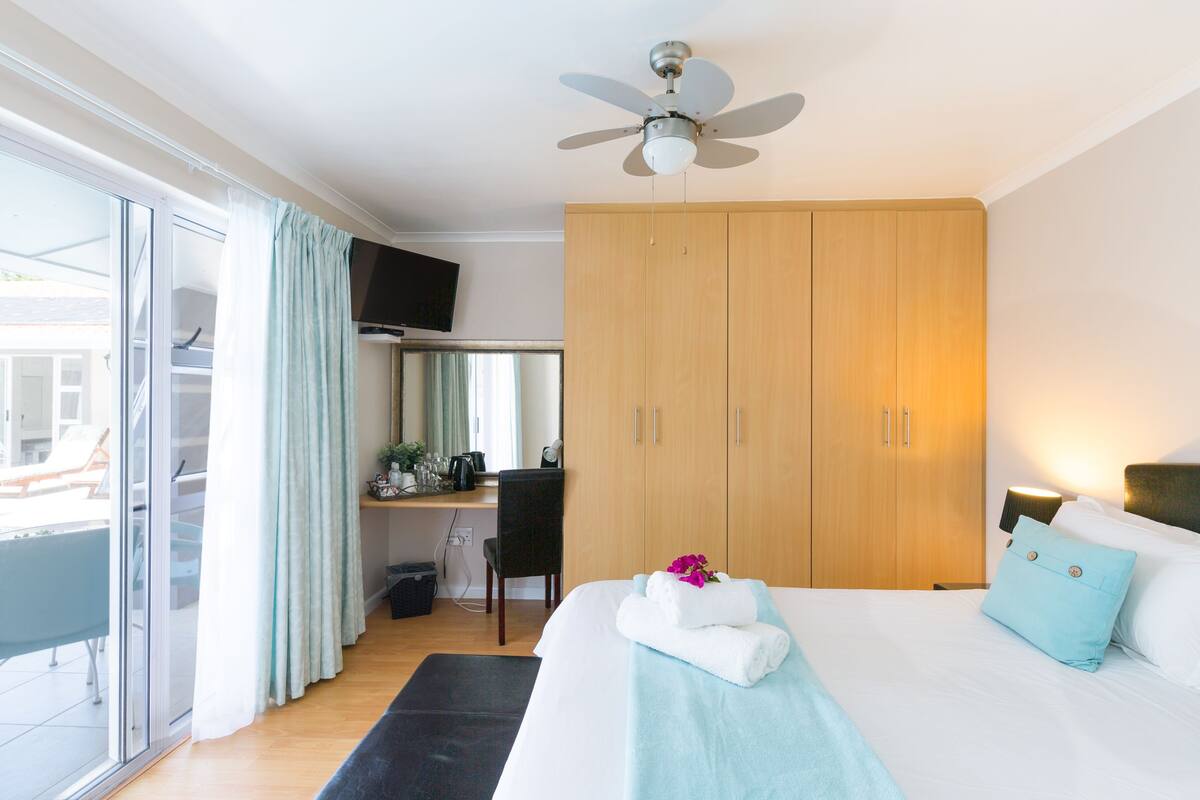 Quarto de hotel com uma cama, escrivaninha, guarda-roupa, ventoinha no teto e vista através de uma porta de vidro deslizante.
