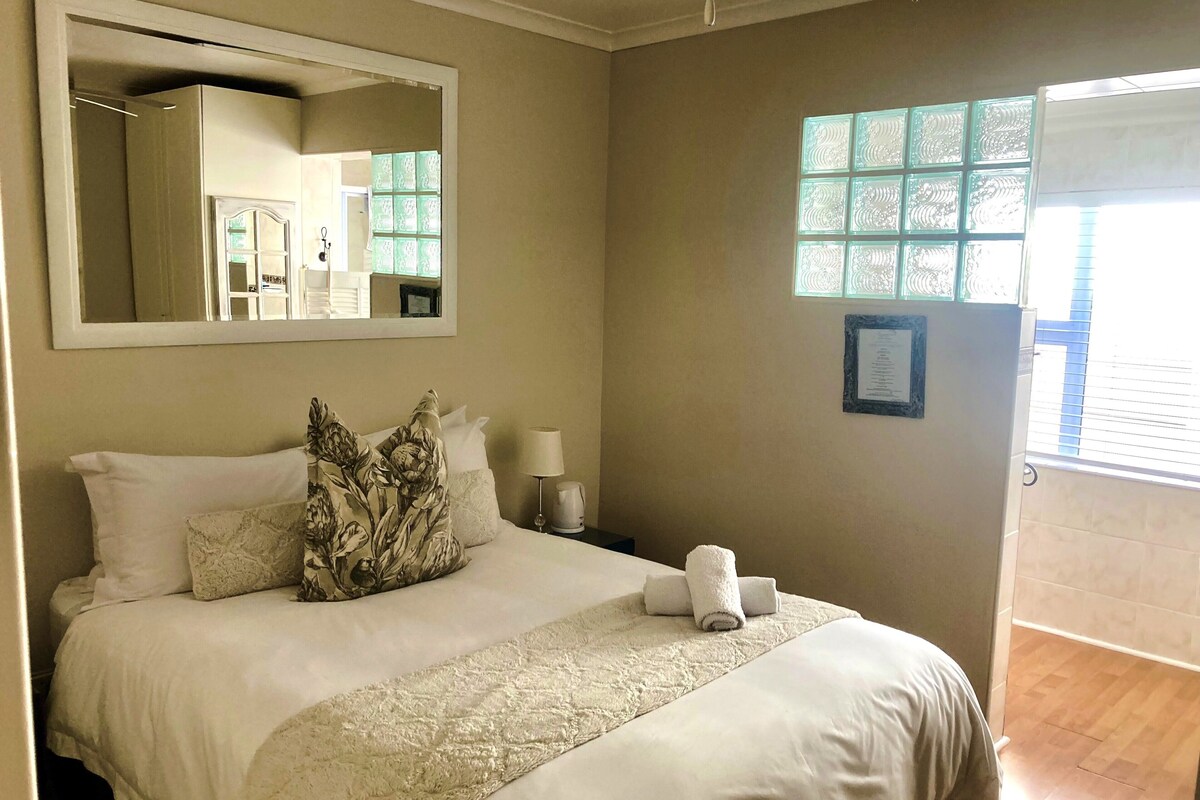 A hotel room with a large bed, a mirror, a nightstand with a lamp, and a window with patterned glass.