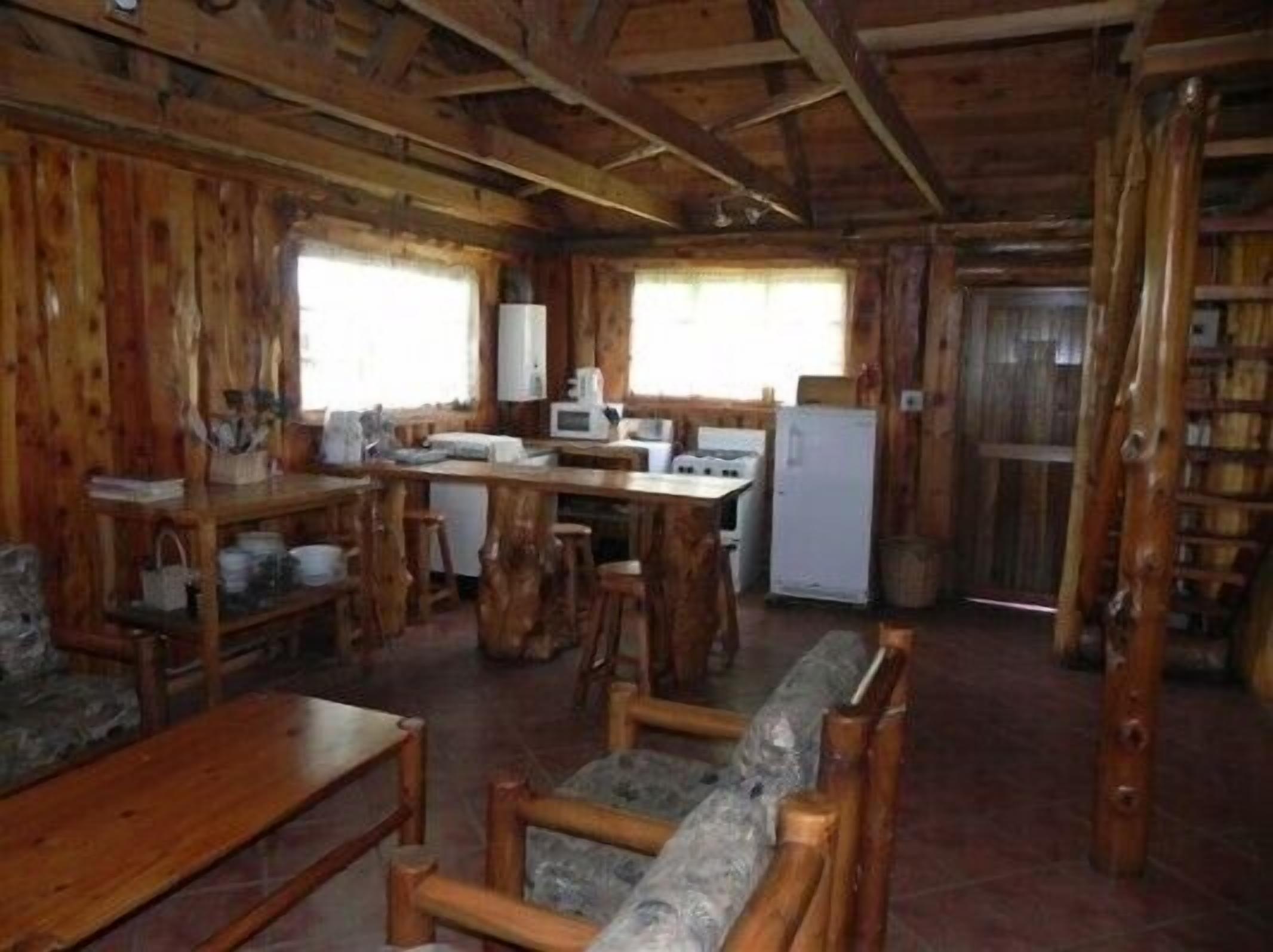 A rustic cabin interior with a wooden dining table, bar stools, a kitchen area with a refrigerator and microwave, and a stone fireplace.