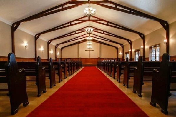 A church interior with pews arranged in rows, a red carpet aisle, wooden beams, and chandeliers.