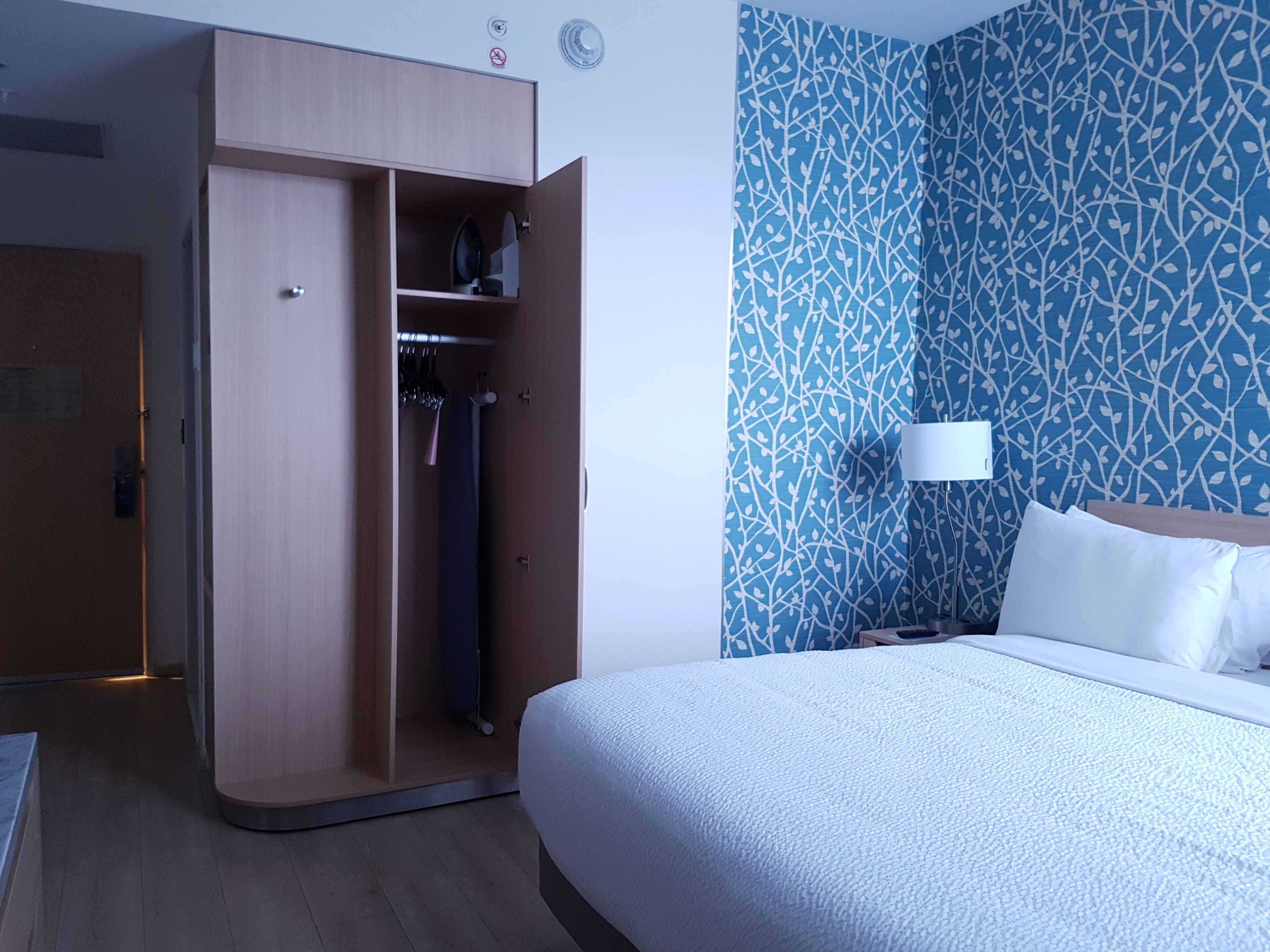 A hotel room with a bed, a wardrobe, a nightstand, and a wall with a leafy pattern.