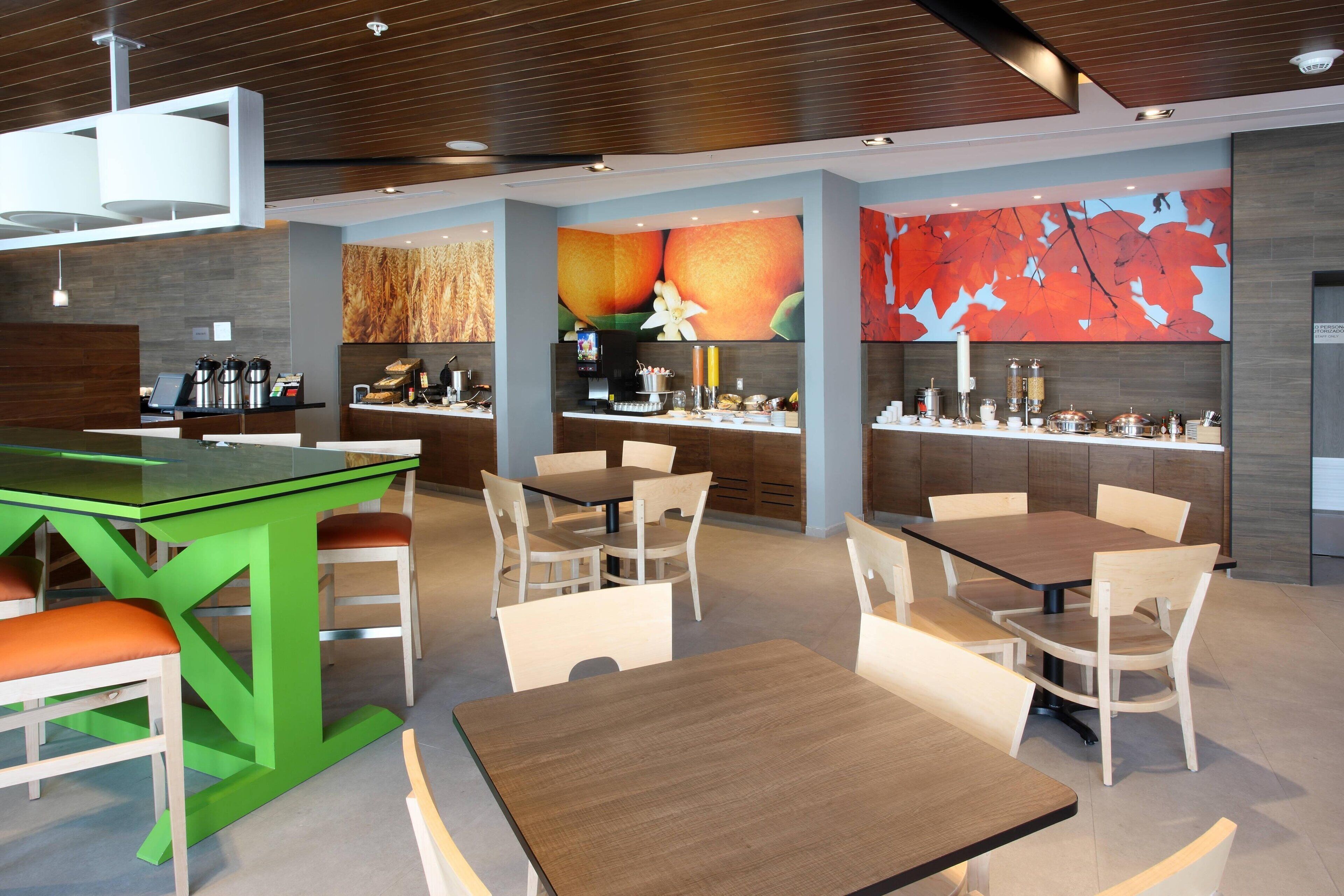 A modern hotel lobby with a green bar, multiple dining tables, and a large fruit-themed mural.