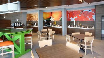 A modern hotel lobby with a green bar, multiple dining tables, and a large fruit-themed mural.