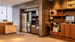A modern hotel lobby with a reception desk, a refrigerator stocked with beverages, a print station, and a wooden wall with a sign that reads "Connect & Print".