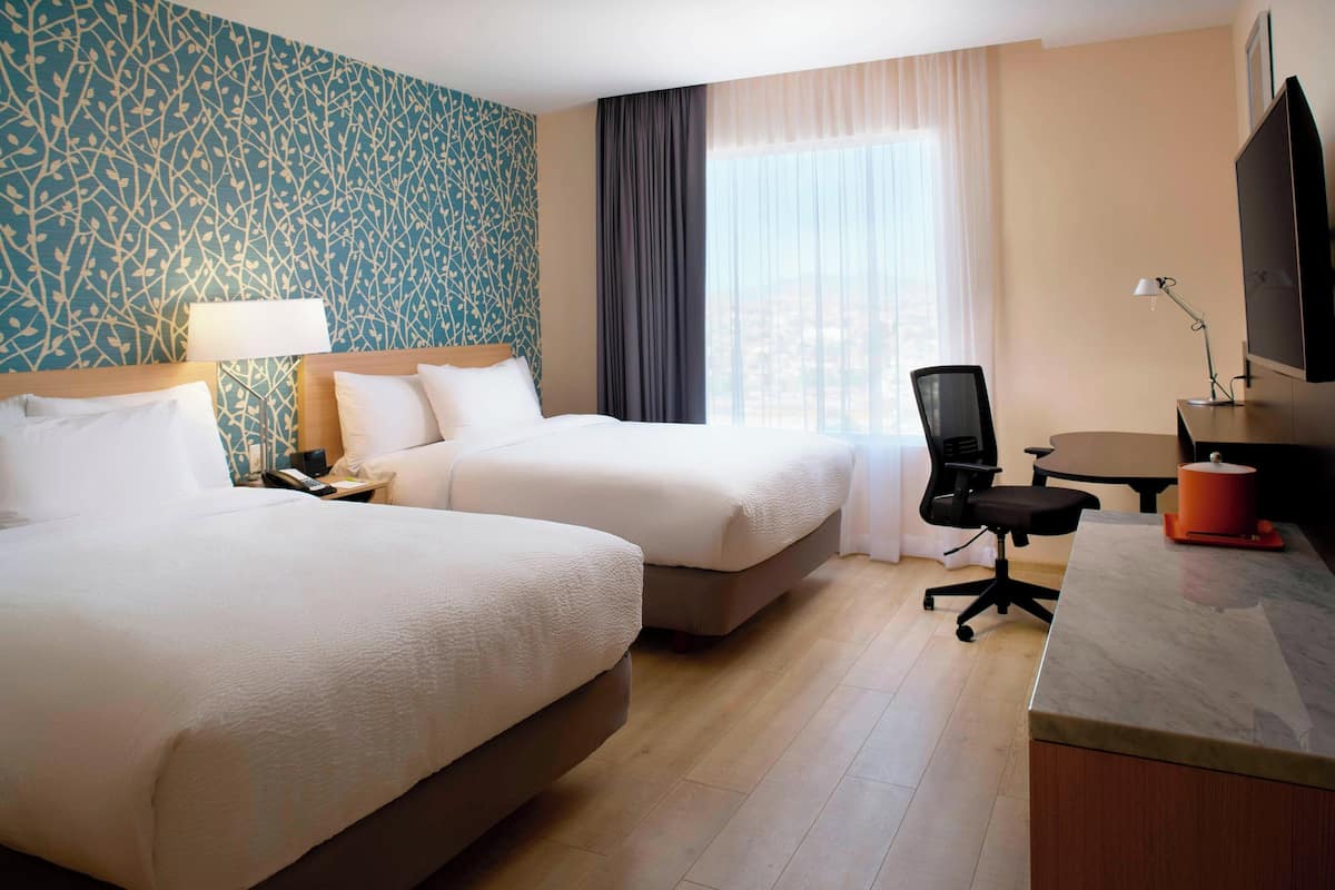 Ein Hotelzimmer mit zwei Betten, einem Schreibtisch, einem Stuhl, einem Fernseher und einem Fenster mit Vorhängen.