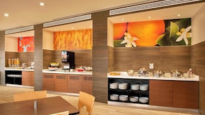 A modern hotel room with a dining area, a kitchen counter with breakfast items, and a large fruit-themed mural on the wall.