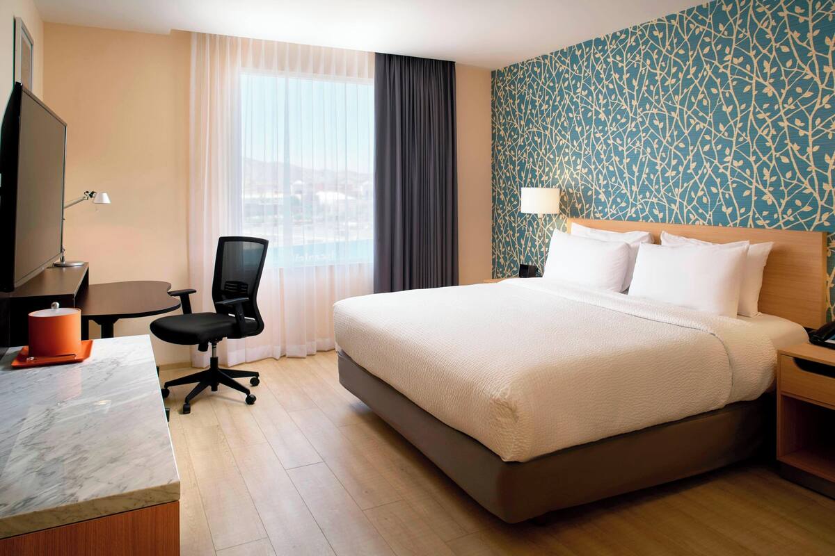 Ein Hotelzimmer mit einem großen Bett, einem Schreibtisch mit Stuhl, einem Fernseher und einem Fenster mit Vorhängen.
