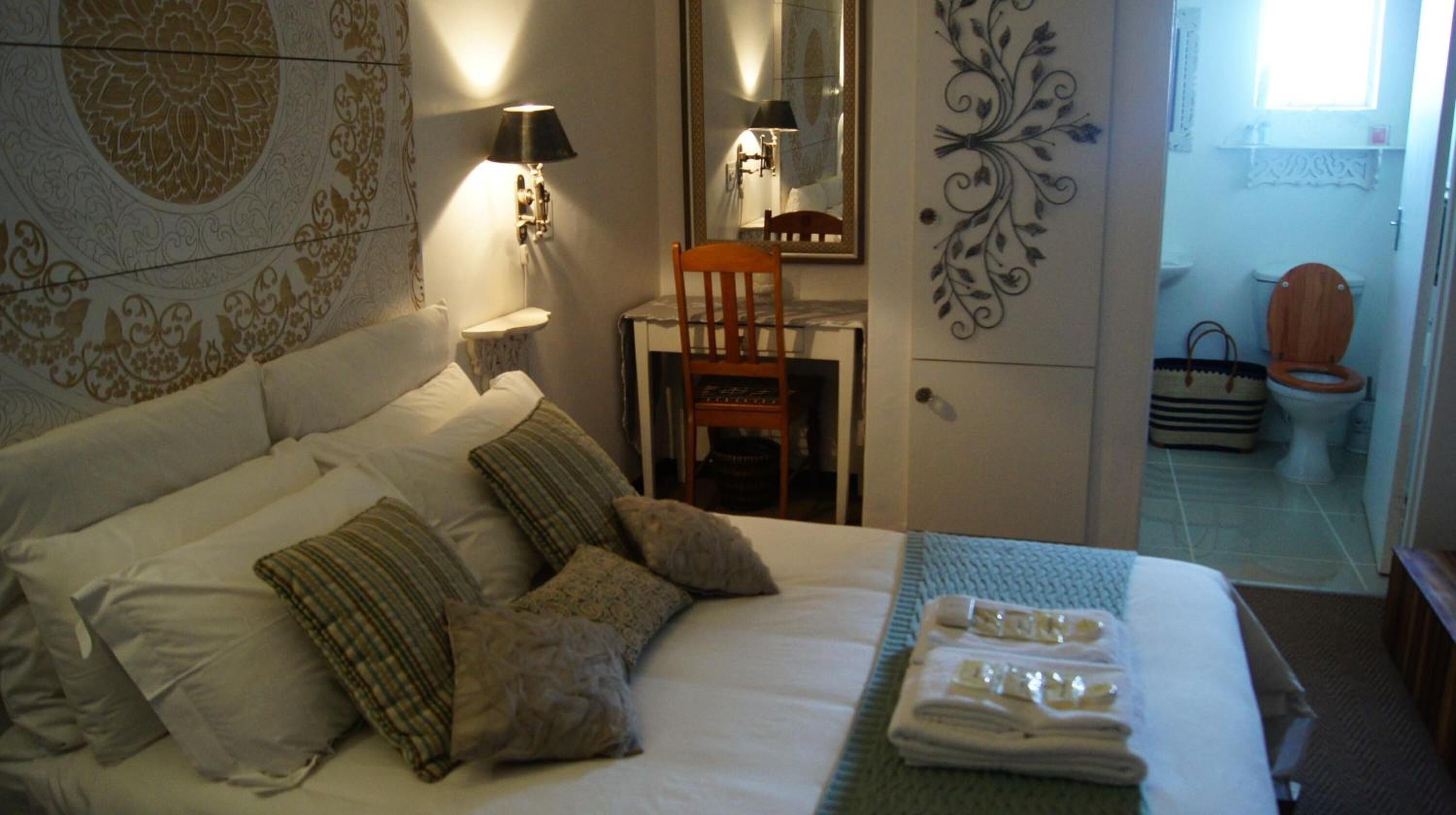 Uma cama bem arrumada com almofadas, uma mesa de cabeceira com abajur, uma cadeira e um banheiro visível através de uma porta aberta.