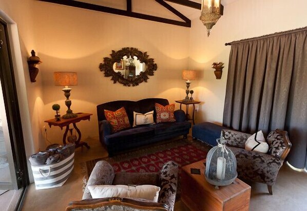 A cozy living room with a dark blue sofa, patterned armchairs, and a wooden coffee table. There is a decorative mirror on the wall and a small side table with a lamp.