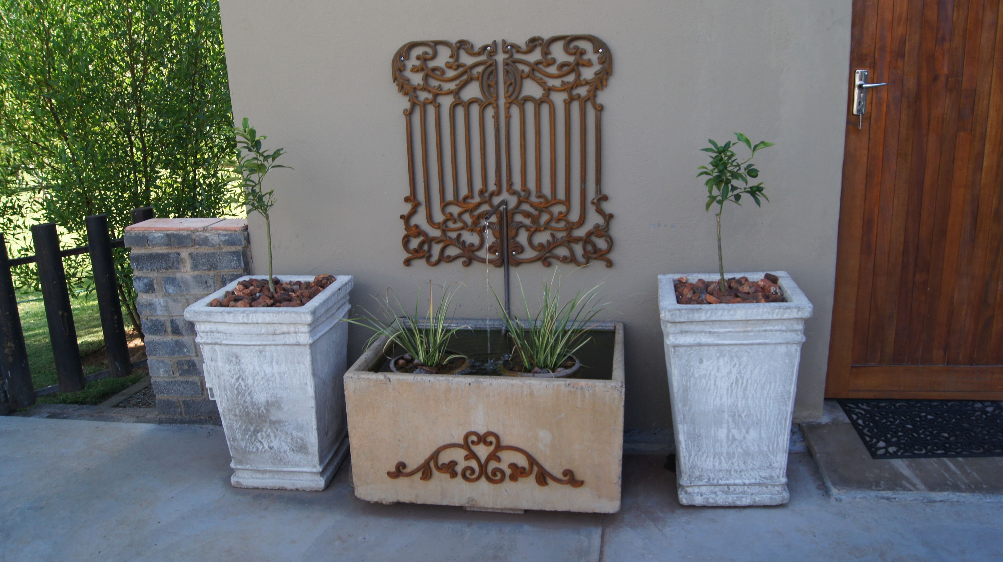 Três vasos retangulares com plantas, um objeto decorativo de metal na parede e uma porta de madeira.