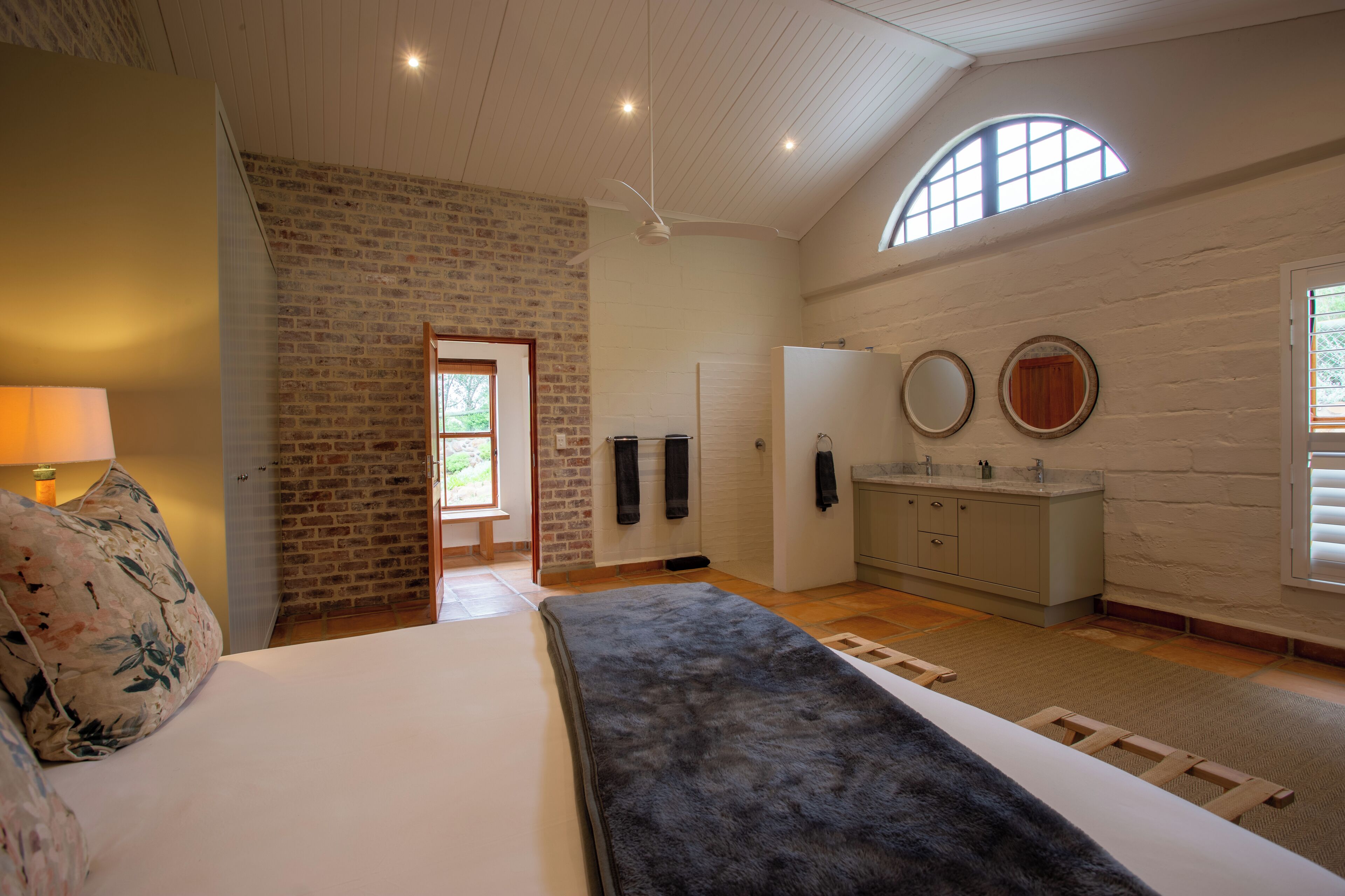A bedroom with a bed, a nightstand, a lamp, a wooden ladder, a brick wall, a window, and a bathroom with a sink and mirror.