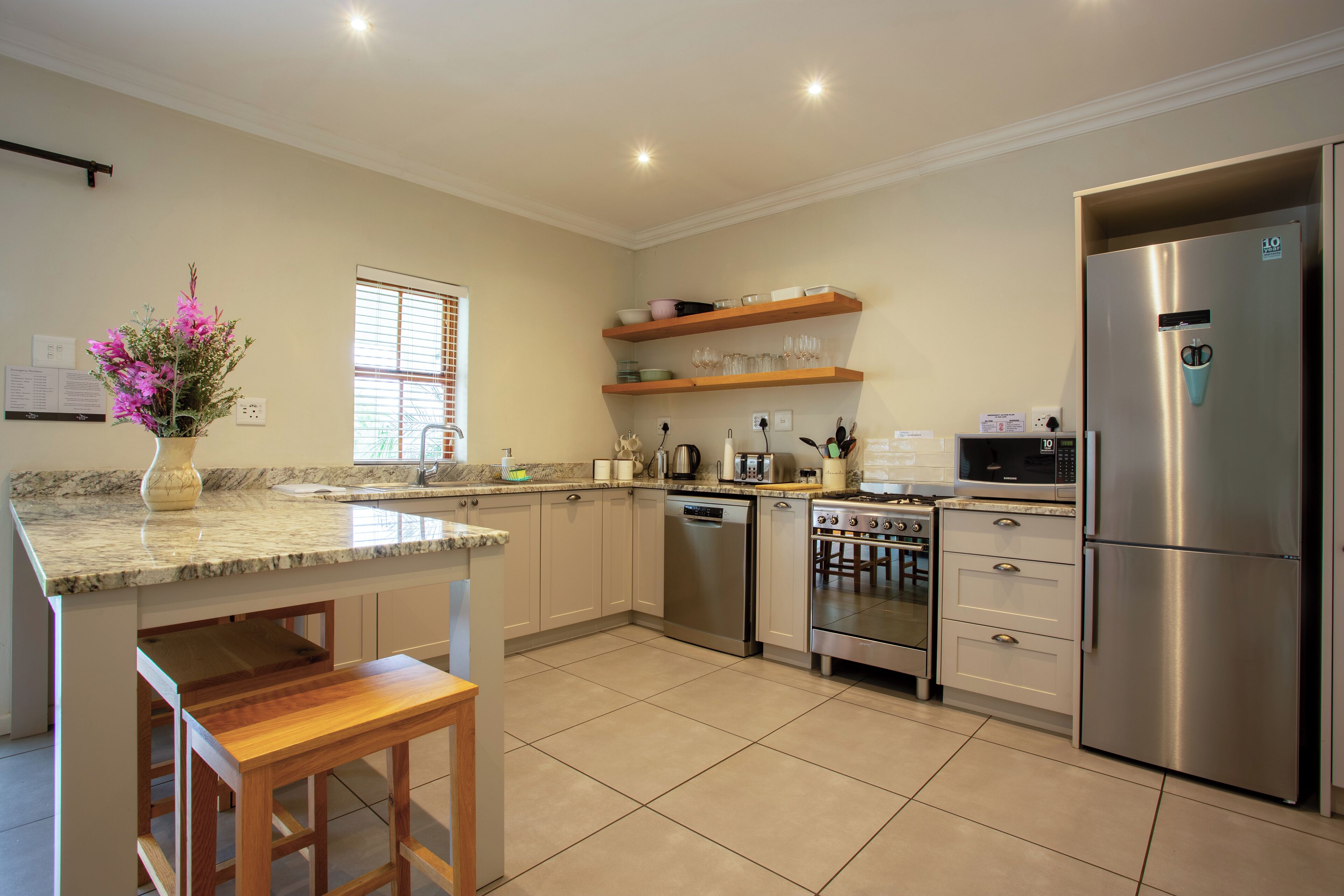 A modern kitchen with a stainless steel refrigerator, built-in oven, microwave, and dishwasher. There is a wooden bench and a vase with flowers on the counter.