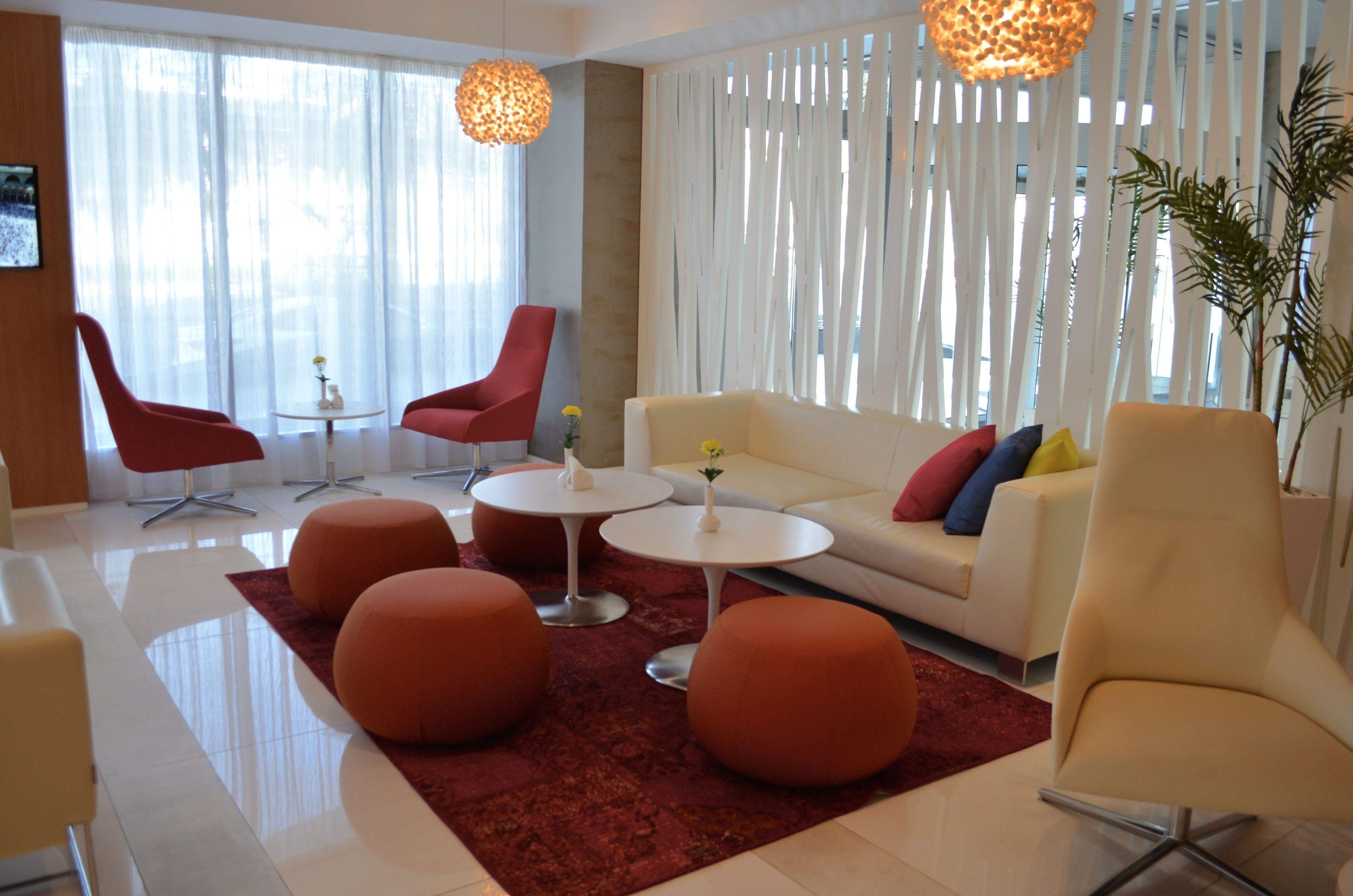 Ein modernes Wohnzimmer mit einem weißen Sofa, roten Sesseln, orangefarbenen Hocker und einem roten Teppich. Zwei weiße Tische mit kleinen Pflanzen darauf.