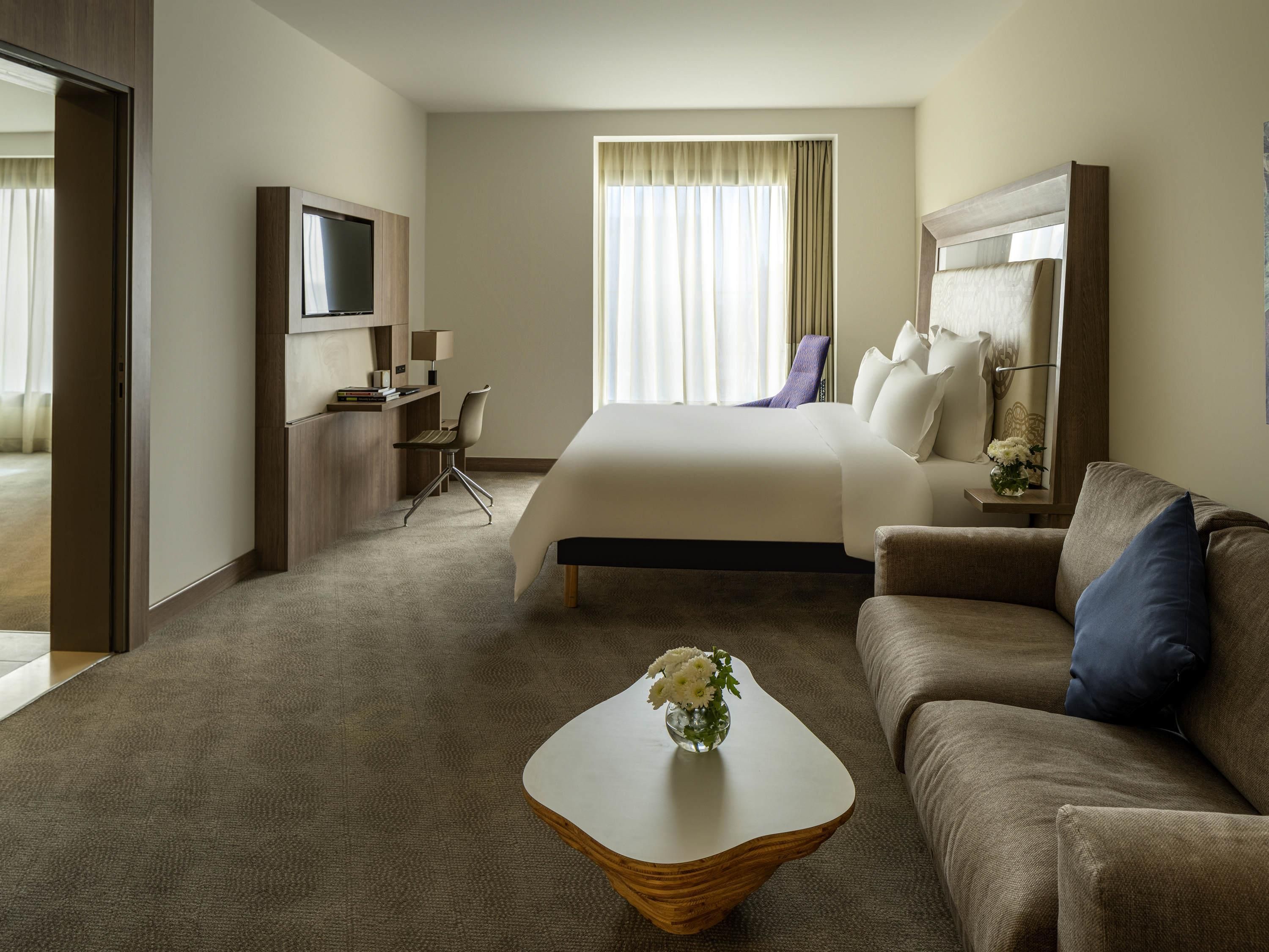 Een hotelkamer met een bed, een bank, een televisie, een bureau en een bijzettafeltje met bloemen.