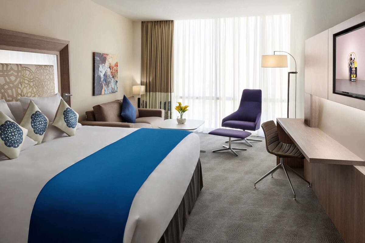 Une chambre d’hôtel avec un grand lit, un bureau, une chaise, une télévision et une fenêtre avec des rideaux.