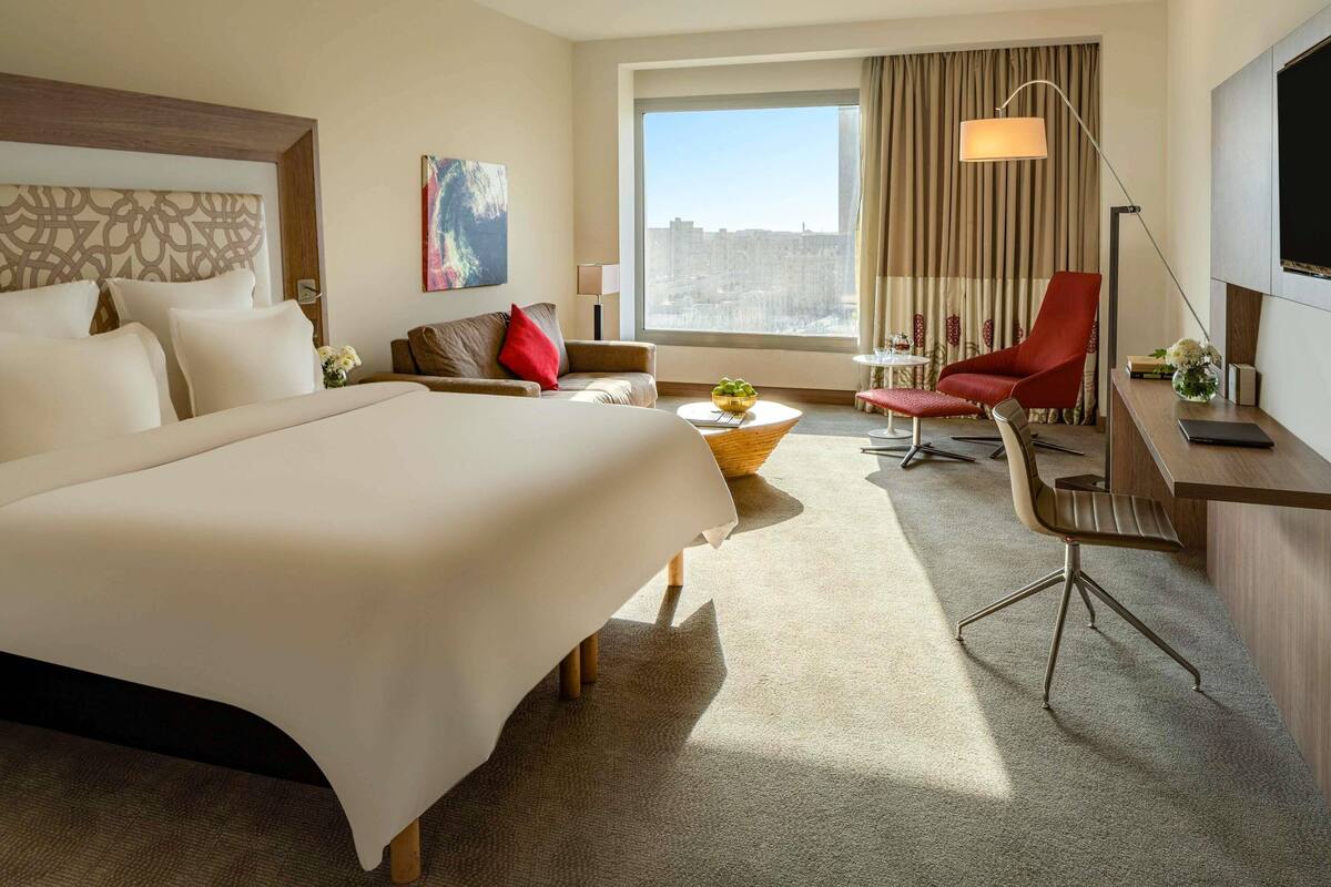 Une chambre d’hôtel avec un grand lit, un bureau avec une chaise, une chaise rouge, une petite table avec un bol, une lampe et un tableau au mur.