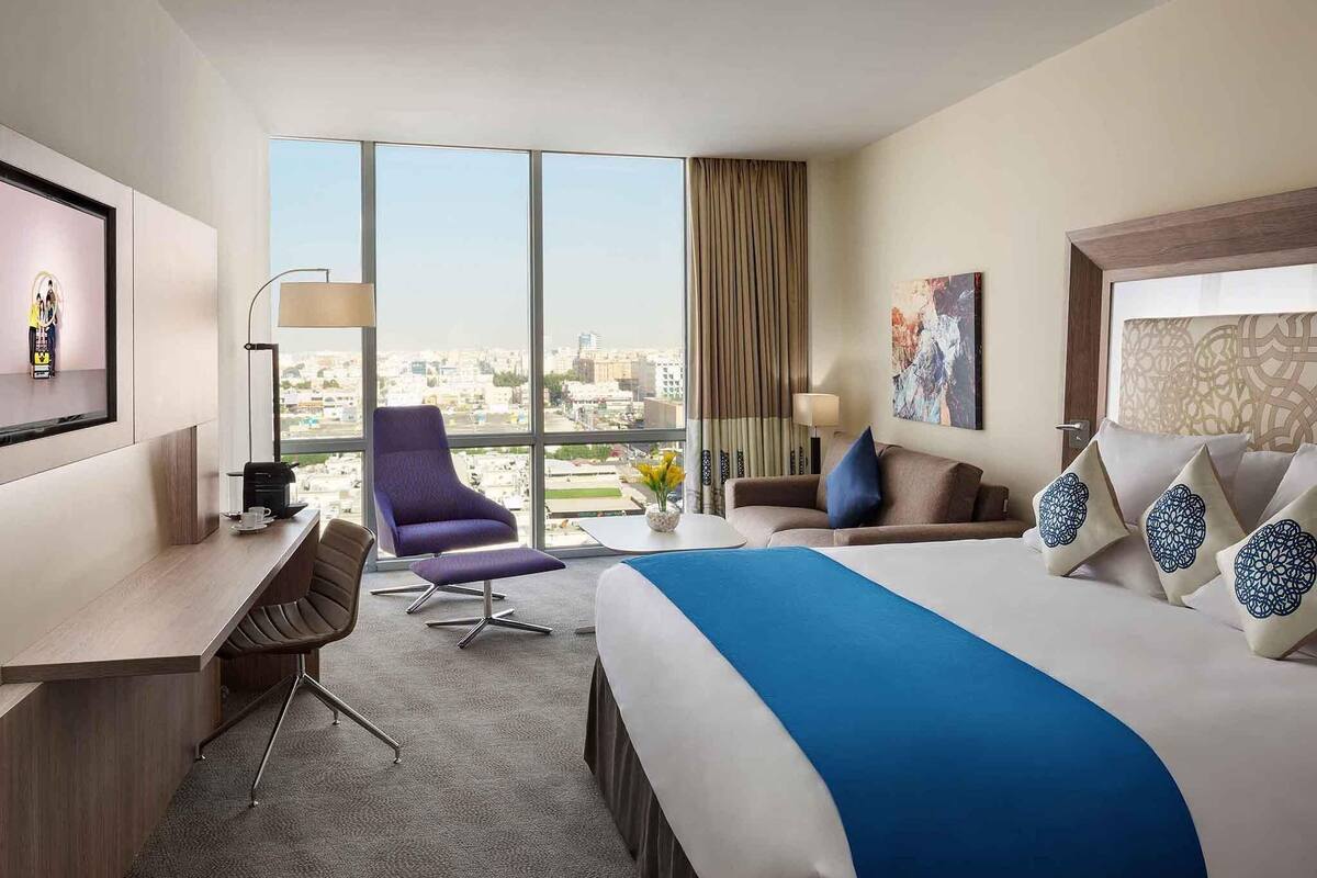 Une chambre d’hôtel avec un grand lit, un bureau, une chaise et une vue sur la ville grâce à de grandes fenêtres.