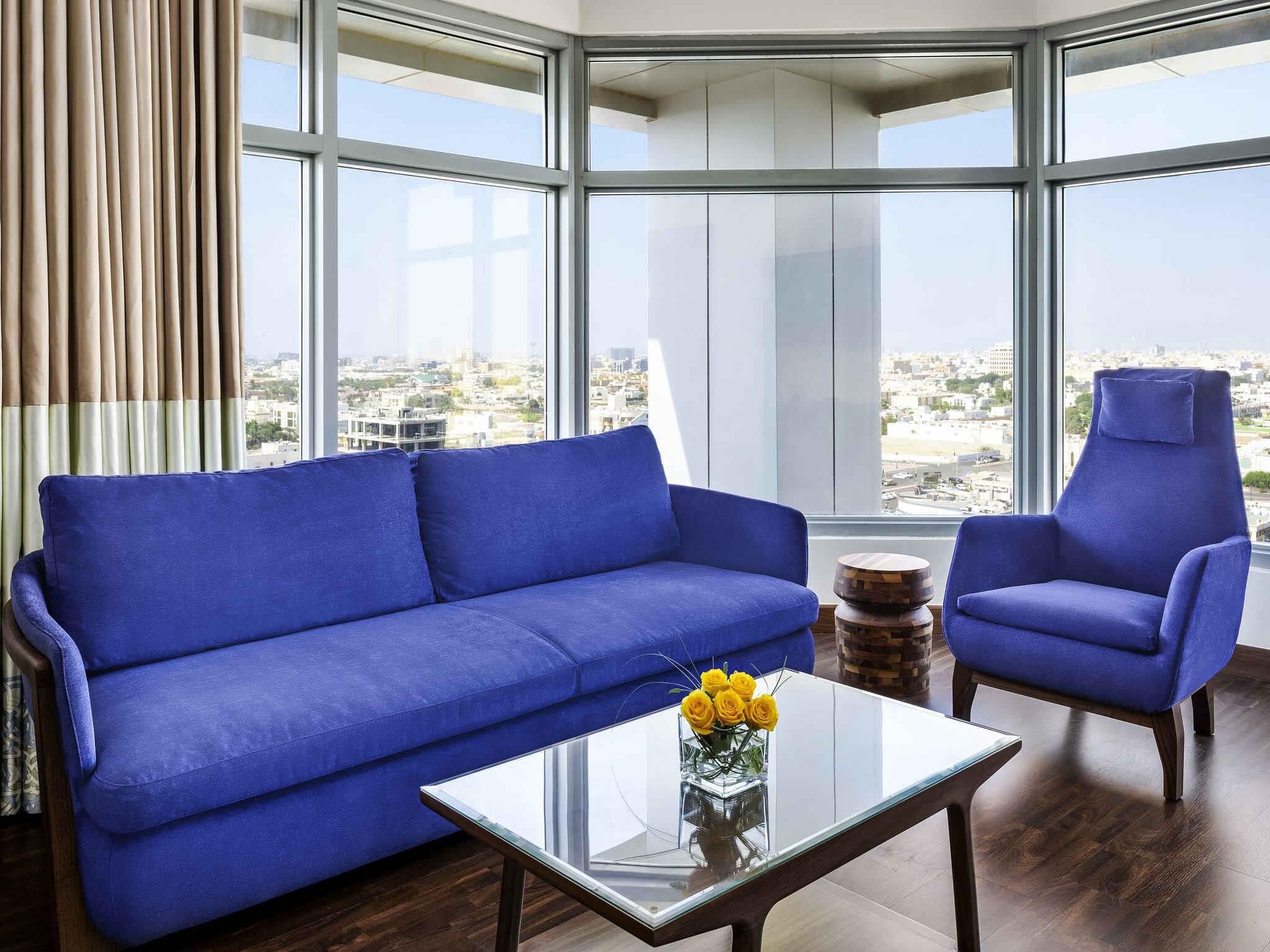 Una sala de estar moderna con un sofá azul, un sillón a juego y una mesa de centro de vidrio con flores amarillas.