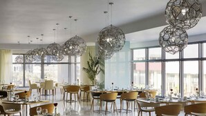 A modern dining area with multiple tables and chairs, large windows, and unique geometric pendant lights. - Novotel Jeddah Tahlia (Jeddah)
