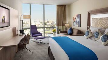 A hotel room with a large bed, a desk, a chair, and a view of the city through large windows.