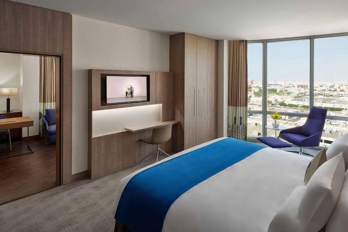 Une chambre d’hôtel équipée d’un lit, d’un bureau, d’une chaise, d’une télévision et d’une grande fenêtre donnant sur la ville.