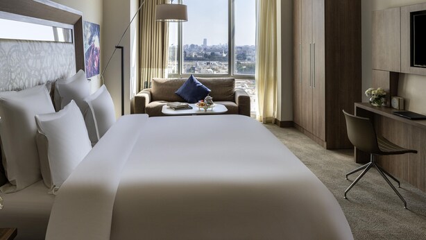 A hotel room with a large bed, a sofa, a desk, a chair, a TV, and a view of the city. - Novotel Jeddah Tahlia (Jeddah)