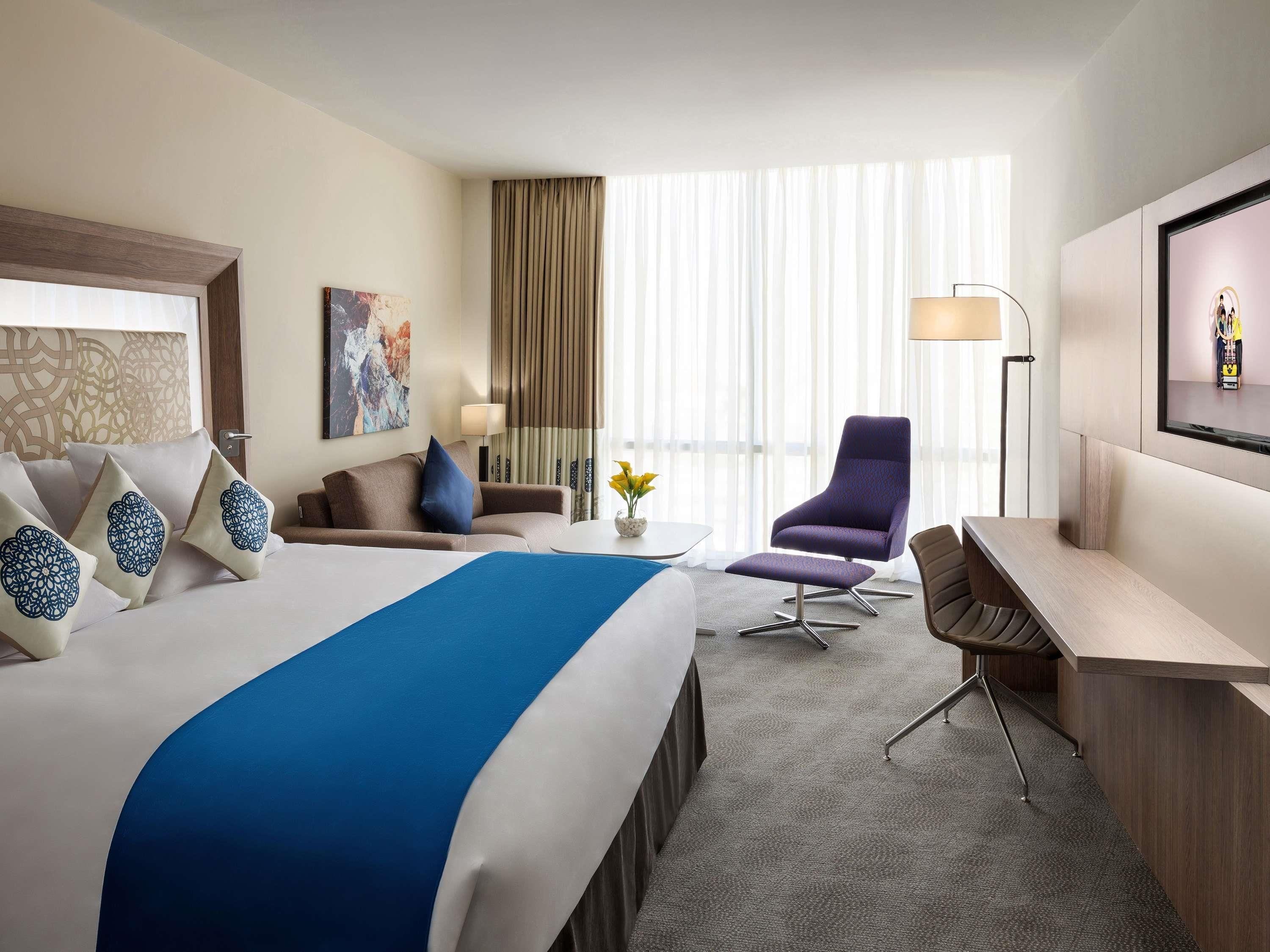 A hotel room with a large bed, a desk, a chair, a TV, and a window with curtains.